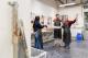 Three people in a workshop wearing aprons and safety gear are being guided through a hands-on task by an instructor.