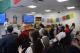 A man speaks to an audience in a classroom decorated with colorful flags and flowers, while people sit and listen attentively.