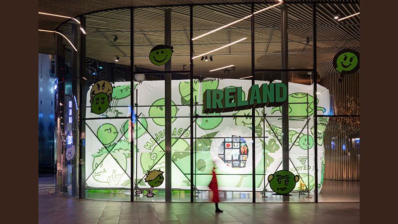 Exterior view of an exhibition installation with a lit inflated tent covered with green logos and graphics