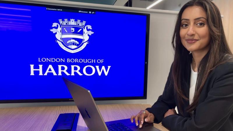 Alumna Kareena Ravaliya posing for a photo in front of her design of the Harrow Logo on a screen behind where she is sitting.