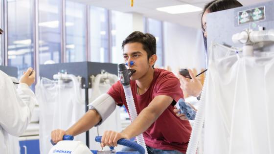 A male athlete cycling on a bicycle whilst someone in a white coat measures their output.