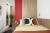 Modern bedroom with a bed featuring patterned pillows and a yellow duvet. A red accent wall and wood paneling behind, with plants and a mirror to the side complete the decor.