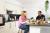 Two friends share a meal in a modern kitchen with white cabinets, black countertops, and green plants. They are smiling and enjoying pastries and orange juice at a kitchen island.