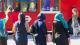 A photograph showing three women who are facing away from the camera. In front of them is a red London bus. The two women on the right are wearing hijabs.