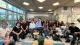 Group photo of approximately 20 people seated and standing in a bright, modern indoor meeting space. The group faces the camera in a semi-circle around small tables. Large windows line the left side of the room, with potted plants placed throughout the space. Overhead ceiling lights illuminate the room, which features lounge-style seating, chairs, and a central coffee table with a plant.