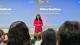 Ritika Wadhwa stands at the front of a lecture theatre addressing an audience seated in rows. Behind the speaker, large presentation screens display a slide featuring the text “Ritika Wadhwa” and information about the author and business leader. The speaker wears a bright pink outfit and stands centre stage, while audience members are visible in the foreground facing the presentation. The room is brightly lit, with the presentation projected across multiple screens at the front of the venue.