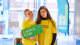 Two female Student Ambassadors wearing yellow t-shirts holding up a green "campus tour" sign