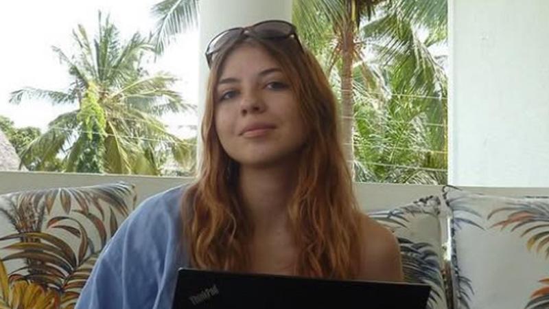Alexandra Bukhareva sitting in front of a laptop smiling. Behind her there are green palm trees.