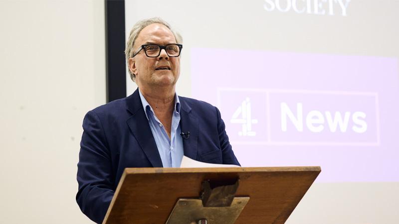 Matt Frei delivering the Hewlett Memorial Lecture at a dais. Behind him is the Channel 4 News logo